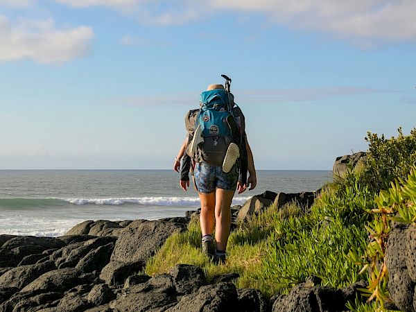 A hiker with a blue backpack stands on rocky shore, looking toward the ocean under a partly cloudy sky.