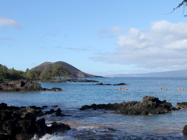 A calm coastal scene with rocky shoreline, blue water, small boats, and a gentle hill in the distance, under a partly cloudy sky.