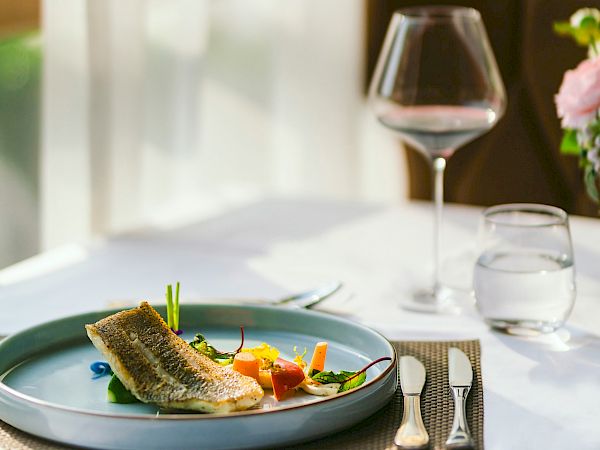 A plated fish dish with vegetables on a blue plate, plus a wine glass, water glass, and a softly lit dining setup by a window.
