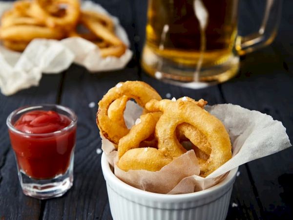 Beer, onion rings, dipping sauce, and a frothy mug on a dark table, with extra rings and salt beside.