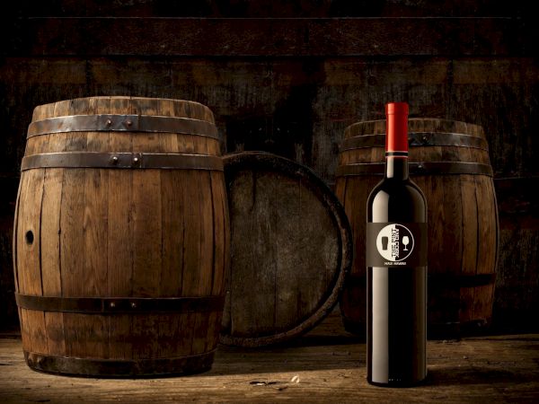 A bottle of red wine with a red foil cap stands beside old wooden barrels in a dim cellar, creating a rustic, vintage ambiance.