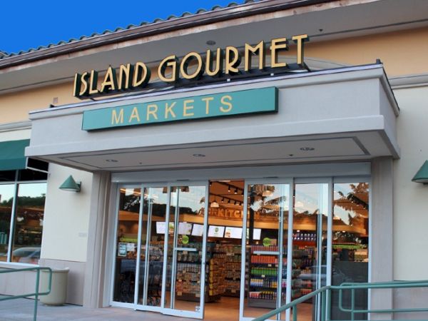 Islands chic Market storefront with glass doors, bright signage &ldquo;ISLAND GOURMET MARKETS,&rdquo; and a sunny, welcoming entrance. End of.