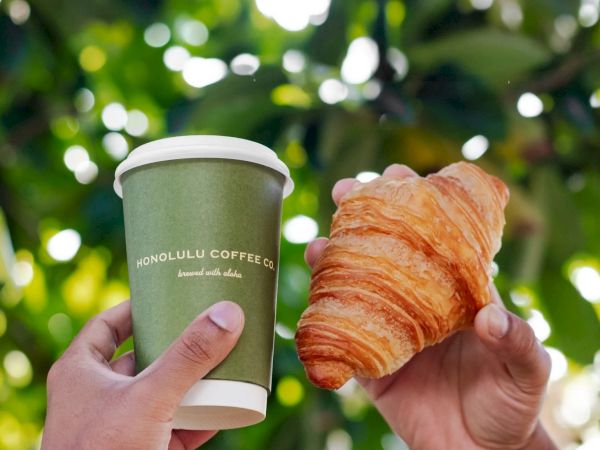 Two hands hold a green coffee cup and a flaky croissant against a leafy, sunlit background. Crisp, fresh, and inviting.
