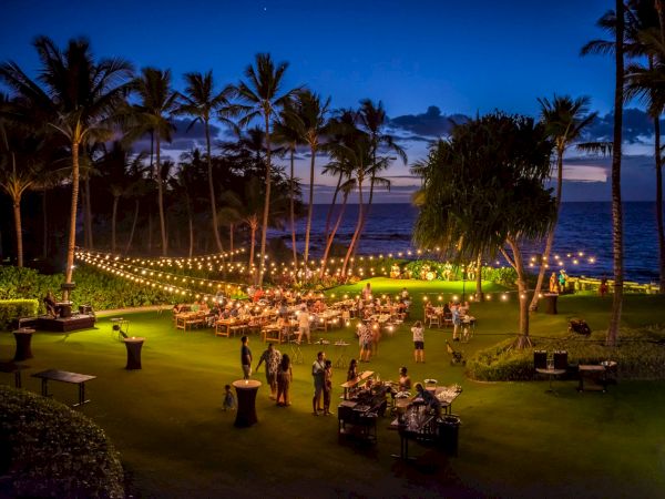 A tropical beachside dinner party at dusk with string lights, palm trees, and guests gathered around tables on a grassy lawn by the ocean.