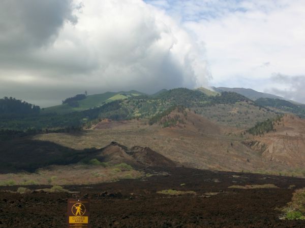 A rocky volcanic landscape with dark lava rocks, rolling hills, and a small warning sign in the foreground, under a cloudy sky.
