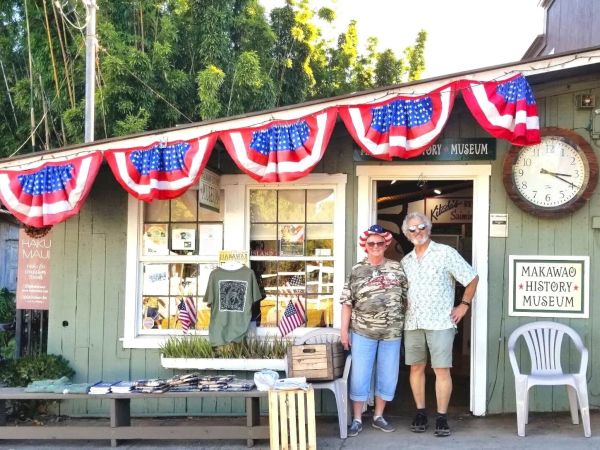 Two people stand in front of a small history museum storefront decorated with American flags and bunting, with souvenirs on a table outside.