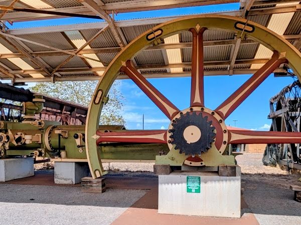 A large vintage wheel artillery-like gear on a stand under a shed, with red spokes and a central blue hub, outdoors with blue sky.