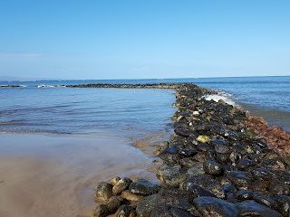 A rocky jetty extends into calm blue sea along a sandy beach under a clear sky, with gentle waves and distant horizon.