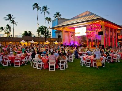 A large outdoor event with many round tables, white chairs, and a lit stage under a pavilion, palm trees in the background, sunset sky.