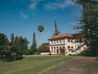 A large, classic two-story mansion with red-tiled roof sits along a curved driveway, surrounded by lush lawns and tall trees on a sunny day.