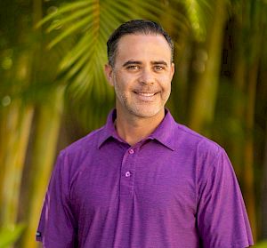 A man in a purple polo shirt stands outdoors, smiling, with palm trees in the background.