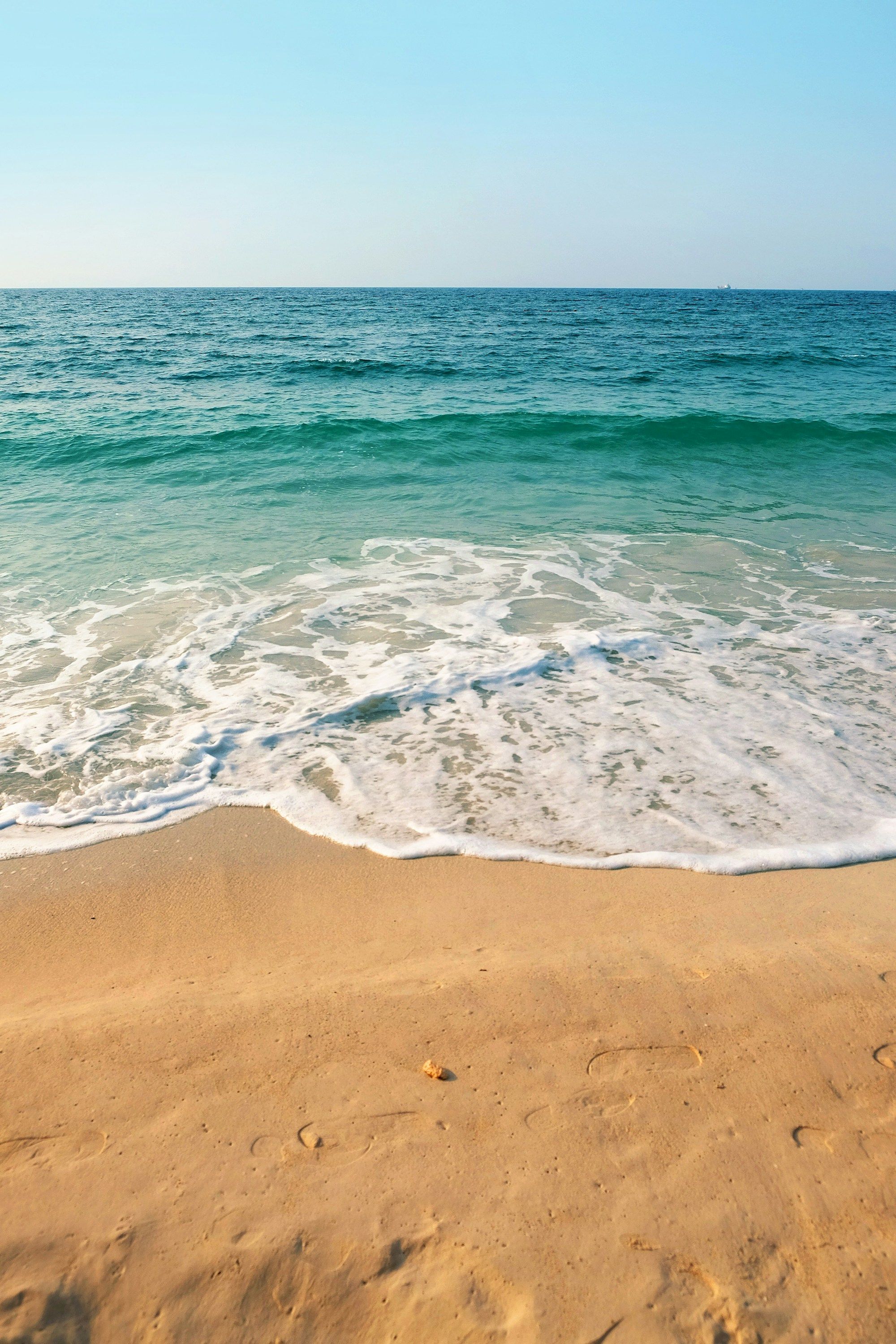 A sunny beach with golden sand, gentle waves washing ashore, and a calm blue-green sea stretching to a clear horizon, peaceful seaside scene.
