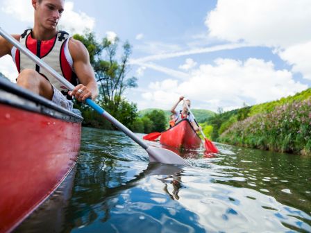 Two people in red kayaks paddling on a calm river, green banks, clear sky, and reflections on the water.