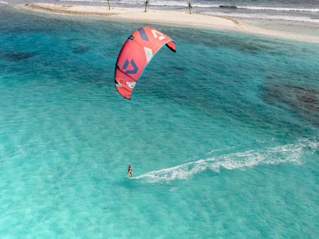 Kite surfer glides over bright turquoise water near a sandy beach, with a red sail catching the wind as the shore and hills appear in the distance.