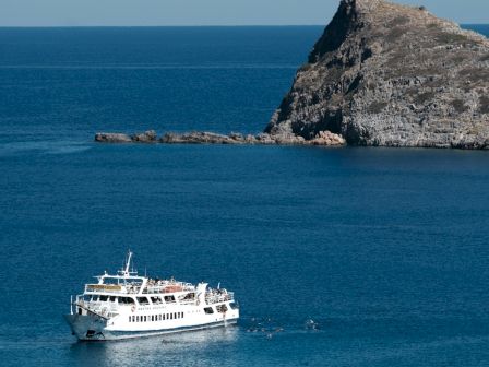 A white passenger boat sails near a rugged rocky outcrop in a calm blue sea, with distant horizon and some foreground shrubbery.