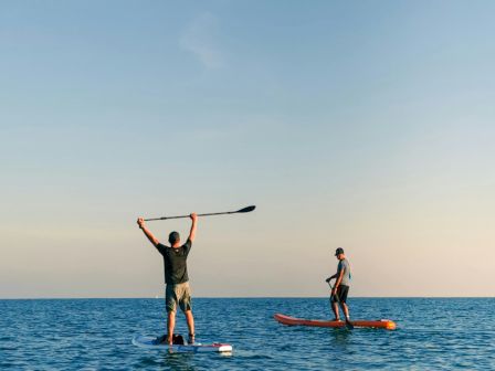 Two people stand on separate boards in calm sea; one raises a paddle overhead while the other watches in the distance, orange sunset sky.