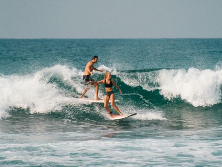 Two people surfing a small to medium wave together, riding close to a breaking crest near the shoreline.