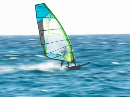 A windsurfer glides across blue waves with a bright, turquoise-green sail, carving through spray under a clear sky.