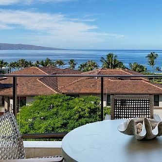 A sunny resort pool area with lounge chairs, a wooden deck, palm trees, and a view of the ocean under a clear blue sky.