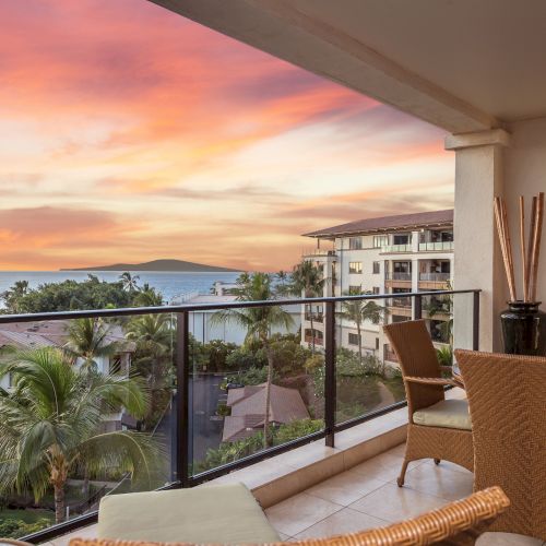 A sunny balcony with a view of palm trees and rooftops, creamy walls, and comfy seating overlooking a warm, scenic landscape.