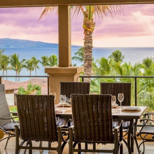 A dining set on a shaded patio overlooking a green lawn and palm trees, with a distant water view and warm sunset light.