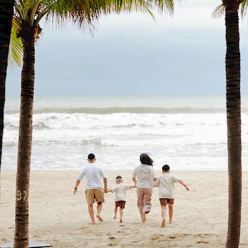 Three kids walk along a sunny beach between palm trees toward the ocean.