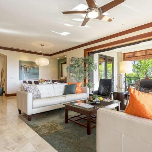 A bright living room with a ceiling fan, large windows, a cozy gray sofa, orange pillows, and a glass coffee table set against a sunny, airy backdrop.