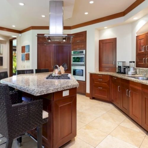 A modern kitchen with warm wood cabinets, a central island, bar seating, and stainless appliances; bright lighting and tiled backsplash.
