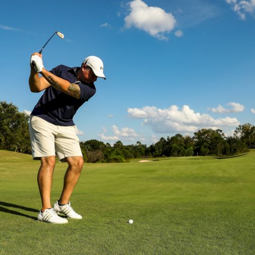 A person wearing a colorful outfit stands on a sunny golf course, holding a club with a blue sky and green hills in the background, smiling.