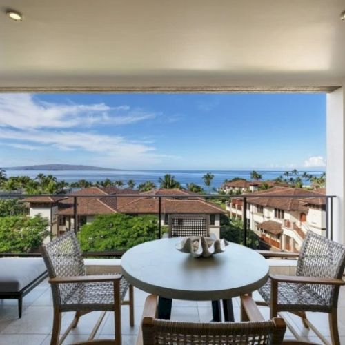 A sunny balcony with a round table and four chairs, overlooking a coastal town and blue water in the distance, bright and inviting.