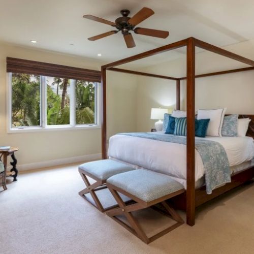 A bright four-poster bedroom with a queen bed, wooden frame, large windows, a ceiling fan, and stylish neutral decor, cozy and airy.