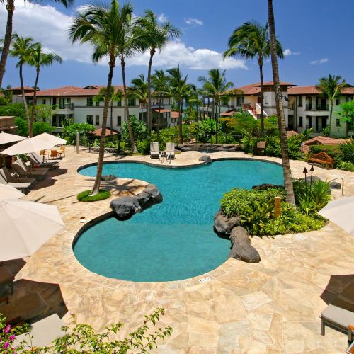A luxurious resort pool with a winding turquoise lagoon, palm trees, tan loungers, rock formations, and a bright blue sky.