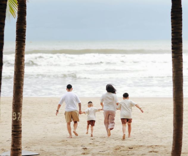 A family walks hand-in-hand along a sandy beach between palm trees toward the waves, enjoying a sunny seaside day.