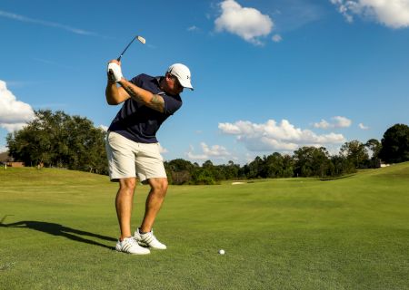 A golfer in a white cap, navy shirt, and light shorts swings a club on a sunny golf course, ball on the tee ready for a drive.