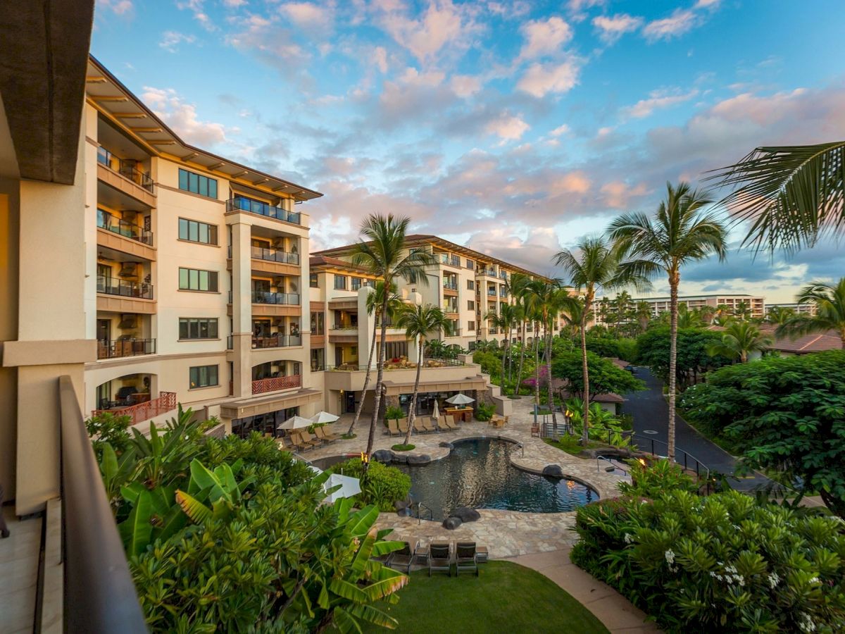 A tropical resort courtyard with palm trees, a pool, lounge chairs, and multi-story beige buildings under a blue sky with scattered clouds.
