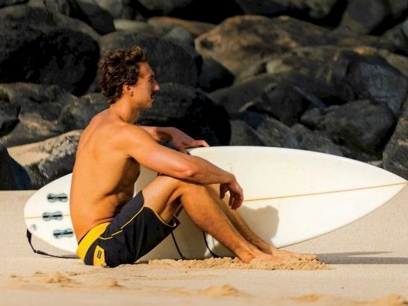 A shirtless man sits on a sandy beach with a surfboard, leaning on rocks and looking out, wearing dark shorts, coastal scene.