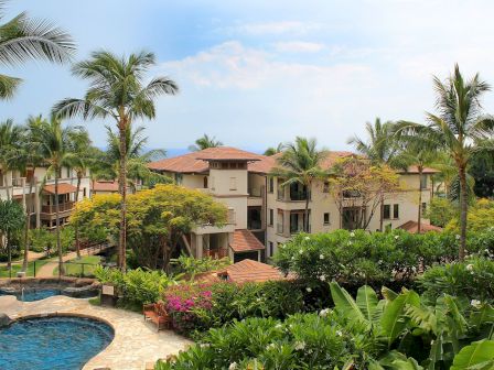 Tropical resort with palm trees, a curved pool, and beige two-story buildings surrounded by lush greenery and flowering plants.