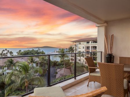 A sunny balcony with wicker chairs and a glass railing overlooks palm trees, a distant island, and a colorful sunset over the sea.