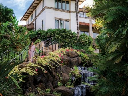 A modern house perched on a lush, tropical hillside with a cascading waterfall, palm trees, and vibrant greenery.